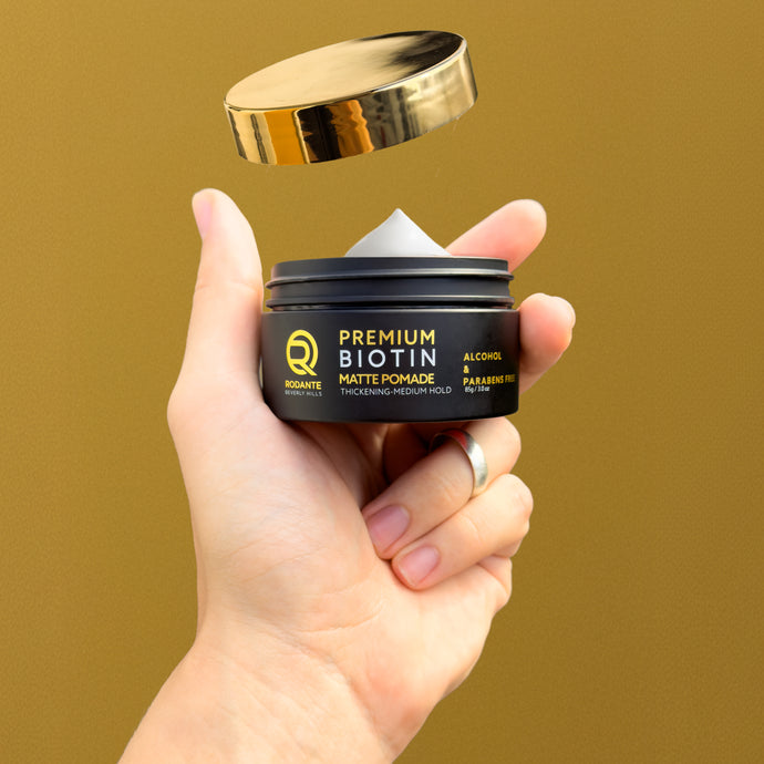 Hand holding a jar of Premium Rodante Biotin Matte Pomade with a gold lid against a brown background